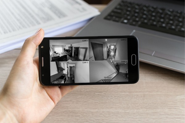 A hand holds a smartphone displaying a grid of four live security camera feeds from different rooms inside a house.