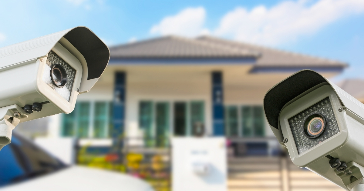 Two outdoor security cameras are prominently featured in the foreground, with a modern house blurred in the background under a blue sky.