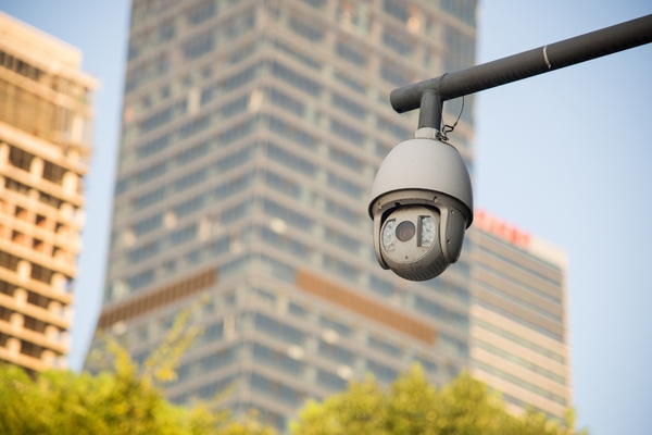 An outdoor dome CCTV camera on a pole with blurred city buildings in the background, showing typical outdoor camera placement.
