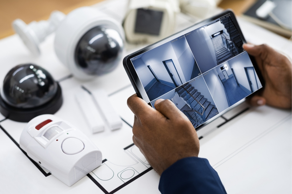 A person monitors multiple security camera feeds on a tablet, surrounded by security devices like cameras and motion sensors.