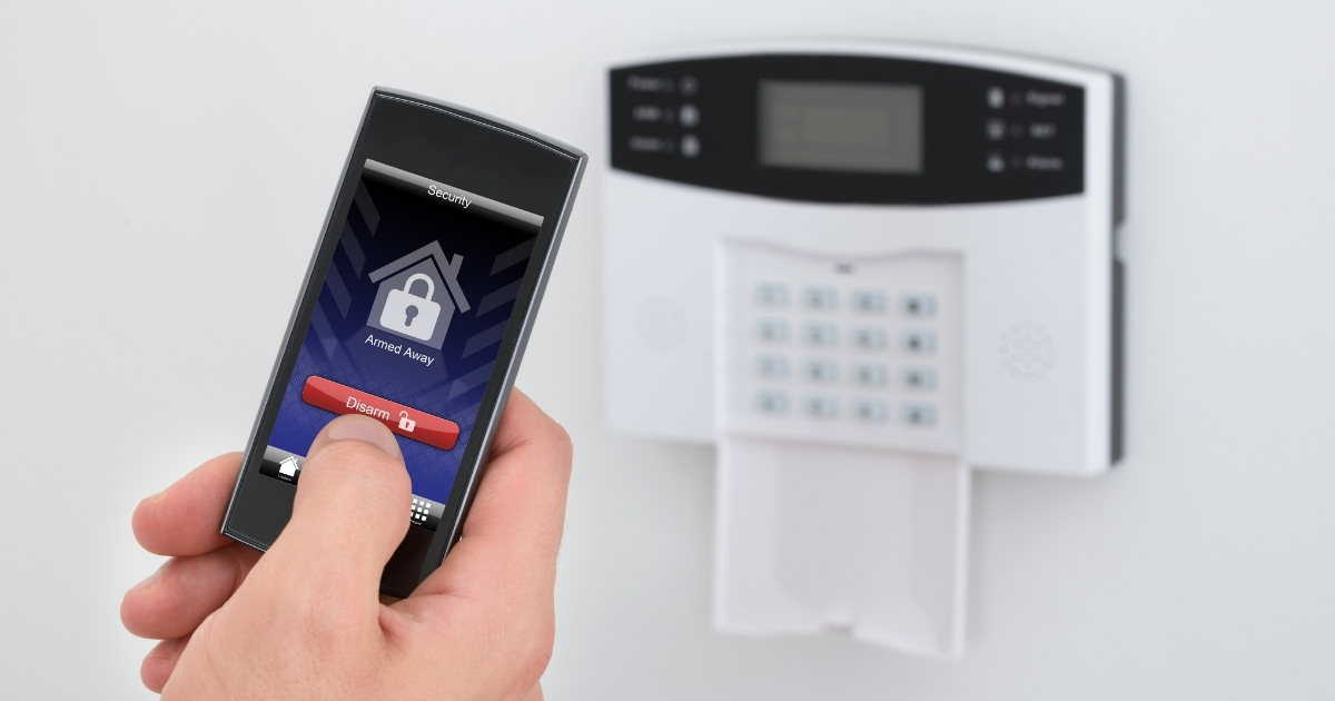 A hand holding a smartphone displaying a security app with an "Armed Away" status, next to a traditional alarm keypad on a wall.