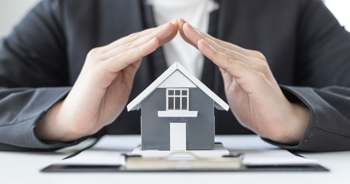Hands forming a protective shape over a miniature house model, symbolizing security and protection.